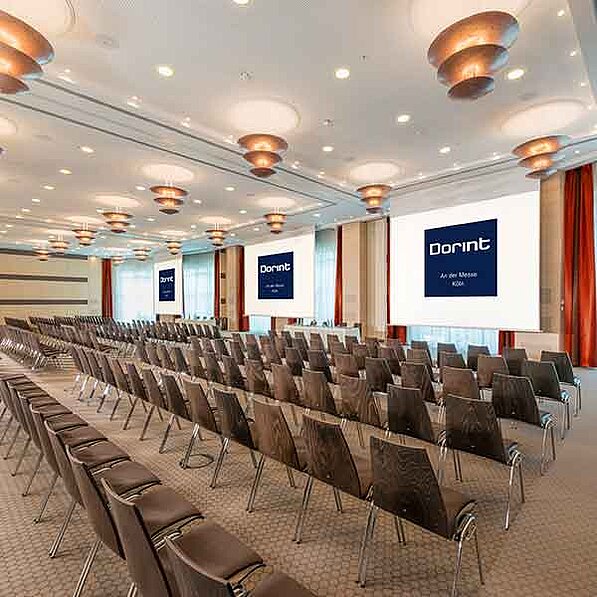 Der Raum ist geräumig und elegant mit einer hohen Decke und moderner Beleuchtung ausgestattet. Reihenweise stehen braune Konferenzstühle auf einem hellen Teppichboden, gerichtet auf drei große Projektionsleinwände an der gegenüberliegenden Wand. Auf den Leinwänden ist das Logo von „Dorint Ammesse Köln“ zu sehen. Die Wände sind hell, und an den Seiten hängen rote Vorhänge. Die Decke ist mit mehreren modernen, runden Lampen ausgestattet, die ein helles, aber nicht grelles Licht spenden. Die Gesamtatmosphäre ist professionell, ruhig und einladend, passend für eine Business-Veranstaltung oder Konferenz. Es sind keine Personen oder Tiere im Bild zu sehen.