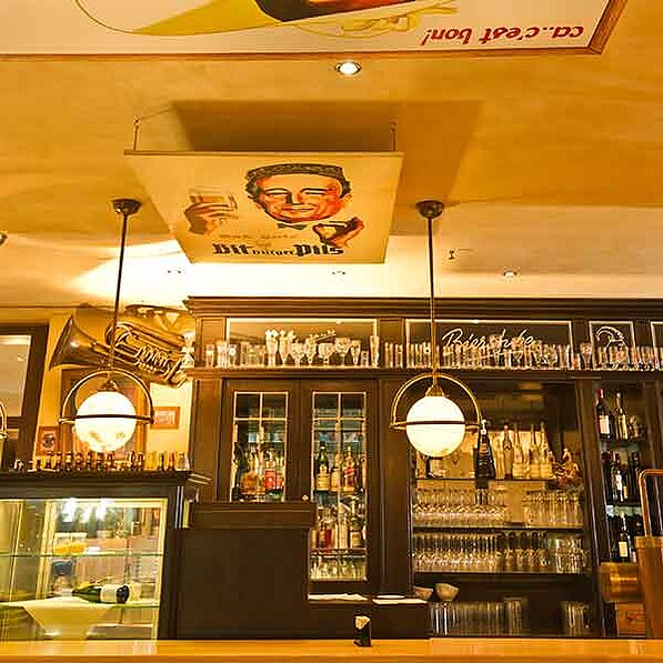 Im Detail zeigt das Bild eine gut beleuchtete Bar mit dunklem Holzmobiliar. An der Decke hängen zwei große, runde Pendelleuchten. Über der Bar ist ein großes, rechteckiges Plakat angebracht, das einen Mann in einem eleganten Anzug zeigt, der ein Glas Bier hält. Das Plakat ist in warmen, goldenen Farbtönen gehalten und weist eine leicht vergilbte Optik auf, was auf einen vintage-Stil hindeutet. Der Mann auf dem Plakat wirkt fröhlich und selbstbewusst. Um das Plakat herum ist die Decke in einem hellen, ockerfarbenen Ton gestrichen.
Hinter der Bar befinden sich Regale, die mit Gläsern verschiedener Größen und Formen sowie einer Auswahl an Flaschen mit alkoholischen Getränken gefüllt sind. Ein Teil der Bar ist verglast, so dass Ein- und Einblicke auf die Getränke möglich sind. Links neben der Bar ist ein Teil einer Theke zu erkennen, auf der einige Flaschen und weitere Gegenstände stehen. Auf der Theke steht auch ein kleines, unklarer Gegenstand, der möglicherweise ein Aschenbecher ist. Ein Teil eines alten, goldenen Tubeninstruments (möglicherweise eine Trompete oder ein anderes Blasinstrument) ist an der Wand neben der Bar befestigt. Die gesamte Atmosphäre wirkt gemütlich, traditionell und einladend, typisch für eine klassische Wirtshaus- oder Kneipenatmosphäre. Die Farben sind warm und erdig, wobei die goldenen Farbtöne des Plakats und der Leuchten den Raum dominieren.