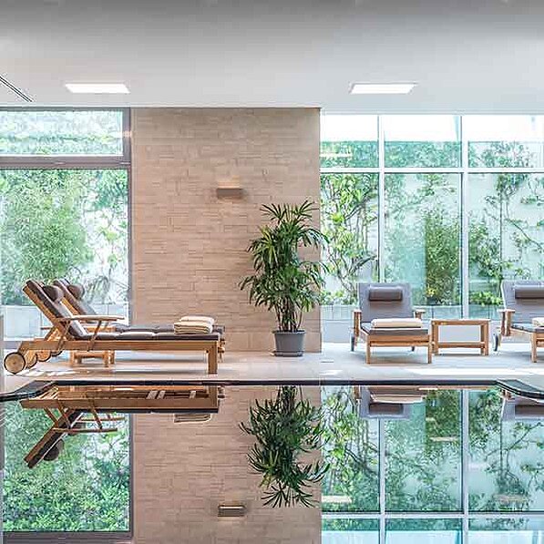 Das Bild zeigt einen modernen, hellen Innenraum, der wohl zu einem Spa oder einem Luxushotel gehört. Der Raum wird von großen Fenstern dominiert, die einen Blick auf üppiges Grün bieten. Im Vordergrund befindet sich ein sauberer, einladender Swimmingpool, dessen Oberfläche wie ein Spiegel die Umgebung reflektiert – die Liegen, Pflanzen und das Grün hinter den Fenstern sind deutlich zu sehen.
An den Pool angrenzend stehen zwei hölzerne Liegestühle aus hellem Holz, die mit weichen Tüchern oder Handtüchern dekoriert sind, einladend und bequem wirken. Auf der gegenüberliegenden Seite des Pools befinden sich zwei weitere, ähnliche, aber einzelne Liegen mit grauen Polstern. Zwischen diesen Einzelliegen steht ein kleiner, ebenfalls aus hellem Holz gefertigter Tisch.
Eine große, grüne Pflanze in einem Topf steht mittig zwischen den beiden Liegen-Gruppen und trägt zur entspannten Atmosphäre bei. Die Wände des Raumes sind mit hellen, sandfarbenen Steinen verkleidet, die eine warme und ruhige Stimmung erzeugen. Die Decke ist weiß und mit eingelassenen Deckenleuchten ausgestattet, die für eine gleichmäßige Ausleuchtung sorgen. Die gesamte Szenerie strahlt Ruhe, Luxus und Entspannung aus. Die Farben sind dezent und natürlich, die Materialien hochwertig und die Atmosphäre insgesamt sehr beruhigend.