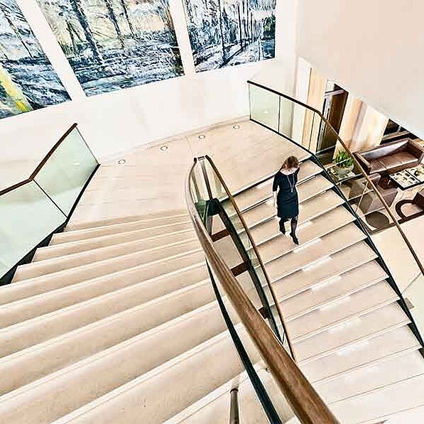 Das Bild zeigt eine elegante, geschwungene Treppe aus hellem Stein, die sich von oben nach unten erstreckt. Die Treppe ist mit einem modernen, gläsernen Geländer versehen, das aus Metall und Holz besteht. Eine Frau in einem dunklen, knielangen Kleid und schwarzen Schuhen geht die Treppe hinunter. Sie trägt eine Halskette. Die Atmosphäre ist ruhig und still. An der Wand hängen zwei große, moderne Kunstwerke, die das Treppenhaus schmücken. Diese Kunstwerke zeigen abstrakte Landschaftsbilder in Blautönen und scheinen mit Pastellfarben gemalt zu sein. Im Hintergrund ist ein Teil eines modernen, hellen Raumes mit einer dunklen Ledercouch und einem kleinen Tisch zu sehen. Pflanzen sind ebenfalls zu erkennen. Die Gesamtkomposition des Bildes ist ordentlich und elegant, mit einer ruhigen, fast meditativen Atmosphäre. Das Licht scheint mild und diffus, so dass keine harten Schatten entstehen.