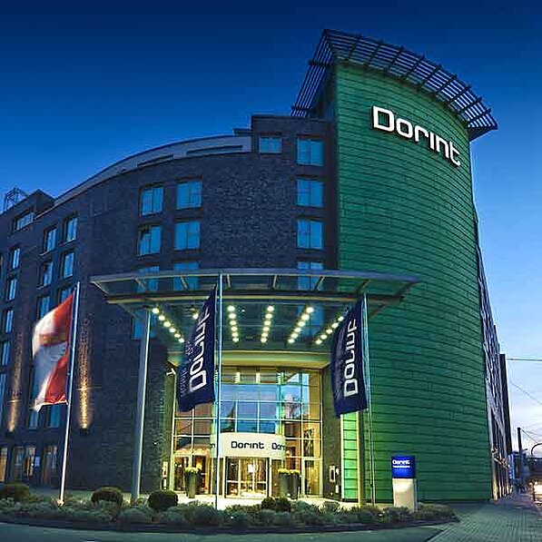 Das Bild zeigt die Außenseite eines modernen Hotels namens Dorint bei Nacht. Das Gebäude ist mehrstöckig und besteht aus dunklem Stein und einem auffälligen grünen Anbau. Der Haupteingang ist von einem großen, gläsernen Vordach überdacht und beleuchtet, unter dem sich die Drehtür zum Hotel befindet. Vor dem Eingang stehen zwei hohe Fahnenmasten mit Unternehmensflaggen. Eine österreichische Flagge ist an einem Mast leicht im Wind wehend zu sehen. Die Hotelfassade ist mit vielen Fenstern versehen, die im Abendlicht hell leuchten. Der Name "Dorint" ist groß und deutlich auf dem grünen Gebäudeteil angebracht. Der umliegende Bereich ist gepflegt und mit niedrigen Pflanzen bewachsen. Im Hintergrund ist ein Teil einer Straße und ein weiterer Gebäudekomplex zu erkennen, die aber unscharf dargestellt sind. Die Atmosphäre ist ruhig und elegant, das Bild strahlt eine gewisse Hochwertigkeit aus. Die Stimmung ist gedämpft und angenehm, typisch für den Abend.