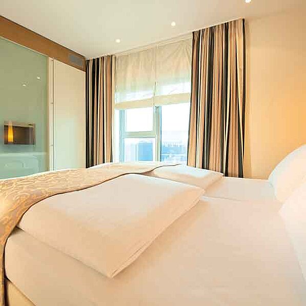 Das Bild zeigt ein Schlafzimmer in einem Hotel oder einer Ferienwohnung. Der Fokus liegt auf einem Doppelbett mit hellbeigefarbener Bettwäsche. Zwei Kissen liegen auf dem Bett. Auf dem Bett liegt eine beigefarbene Tagesdecke mit einem dezenten Muster. Hinter dem Bett befinden sich cremefarbene Vorhänge mit vertikalen, beige- und dunkelbraunen Streifen. Ein hellbeiger, durchsichtiger Vorhang ist über dem Fenster angebracht. Das Fenster selbst zeigt einen Blick auf die Skyline einer Stadt. Der Raum ist hell und freundlich eingerichtet. An der Wand links im Bild ist ein Kleiderschrank mit einer Glastüre zu sehen, in der ein kleiner Flachbildfernseher integriert ist. Die Wände des Zimmers sind in einem warmen, hellen Beige gehalten. Die allgemeine Atmosphäre ist ruhig, entspannend und einladend. Das Bild wirkt sauber und ordentlich und vermittelt ein Gefühl von Luxus und Komfort.